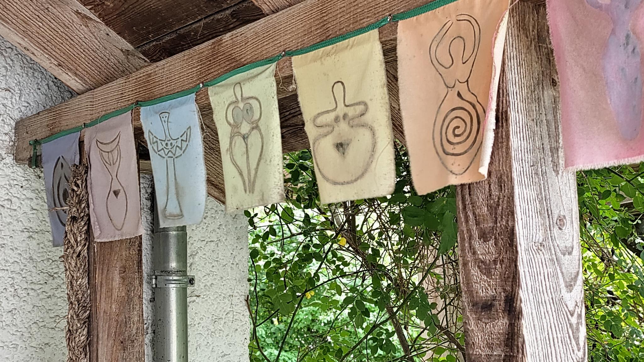 Verschiedene matriarchale Zeichnungen auf kleinen bunten Stoffen, die aneinandergespannt an eine Leine außen am Haupthaus hängen