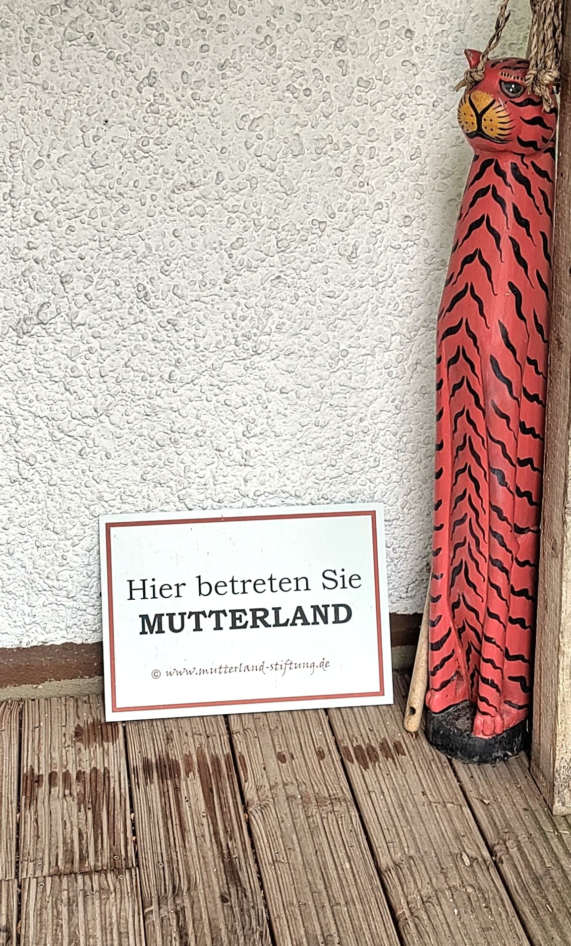 Ein Terassenausschnitt vor dem Haupthaus. Auf dem Holz steht eine lachsfarbene, hölzerne Figur. An ihr lehnt ein Schild auf dem geschrieben steht: Hier betreten sie Mutterland