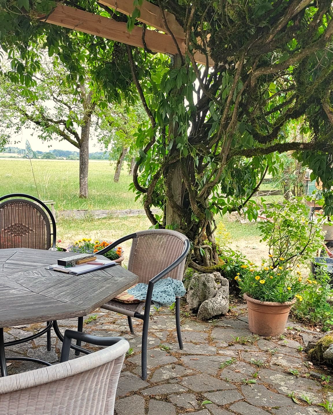 Die Terrasse, runder Tisch mit Stühlen am Haupthaus. Direkt dahinter Blick auf weitläufige Felder