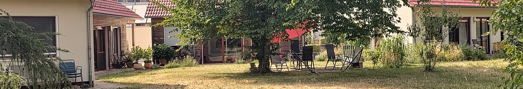 Panoramaaufnahme im Sommer der Tiny Houses in Wennenden. Mann sieht alle vier Häuser, ,manche davon nur seitlich da diese wie eine Wagenburg angeordnet sind. Im Vordergrund sieht man eine Wiese mit Baum und daneben einen Gartentisch mit mehreren Stühlen.